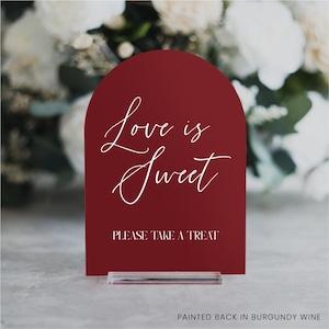 May include: A burgundy-colored sign with an arched top, featuring the words "Love is Sweet" in elegant white script. Below, it reads "PLEASE TAKE A TREAT." The sign is mounted on a clear acrylic base, with a blurred floral background.