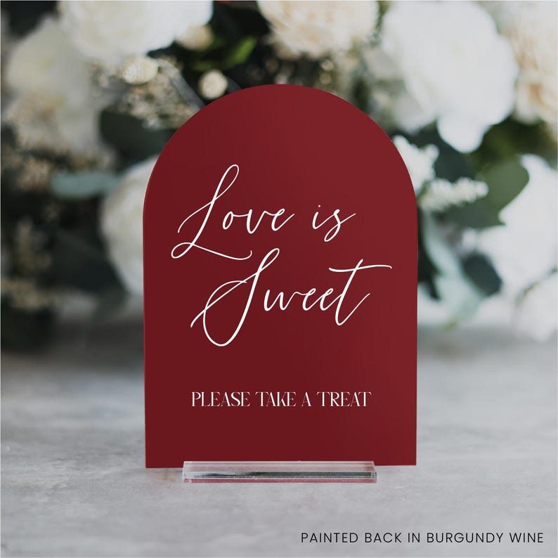 May include: A burgundy-colored sign with an arched top, featuring the words "Love is Sweet" in elegant white script. Below, it reads "PLEASE TAKE A TREAT." The sign is mounted on a clear acrylic base, with a blurred floral background.