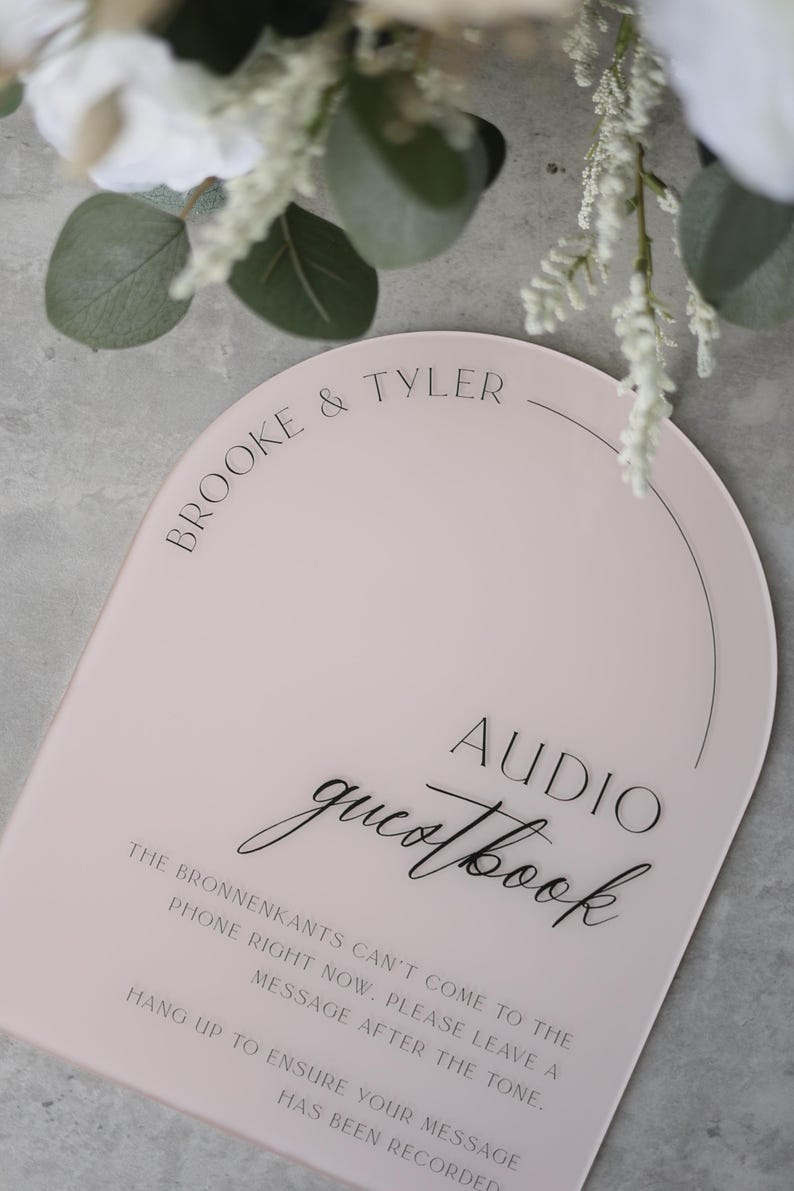 May include: A pink, arched sign with the words "Brooke & Tyler" at the top. Below, it reads "Audio Guestbook" in elegant script. The sign provides instructions for leaving a message.