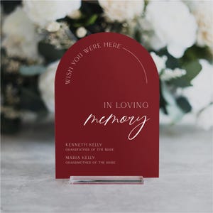 May include: A red acrylic sign with rounded edges and a white inscription that reads "Wish you were here" in a semi-circle at the top. The sign also says "In loving memory" with a handwritten font and lists two names with their relationship to the bride.
