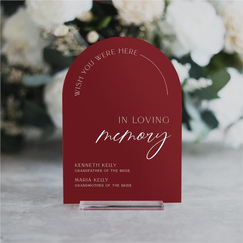 May include: A red acrylic sign with rounded edges and a white inscription that reads "Wish you were here" in a semi-circle at the top. The sign also says "In loving memory" with a handwritten font and lists two names with their relationship to the bride.