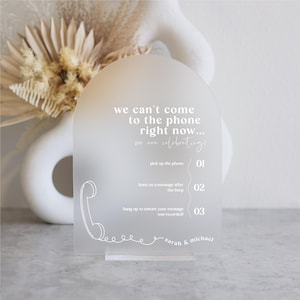 May include: A frosted acrylic sign with a white arch shape and a white phone graphic. The sign reads "We can't come to the phone right now... we are celebrating!" and includes instructions for leaving a message: "Pick up the phone", "Leave us a message after the beep", and "Hang up to ensure your message was recorded!" The sign is signed "Sarah & Michael".