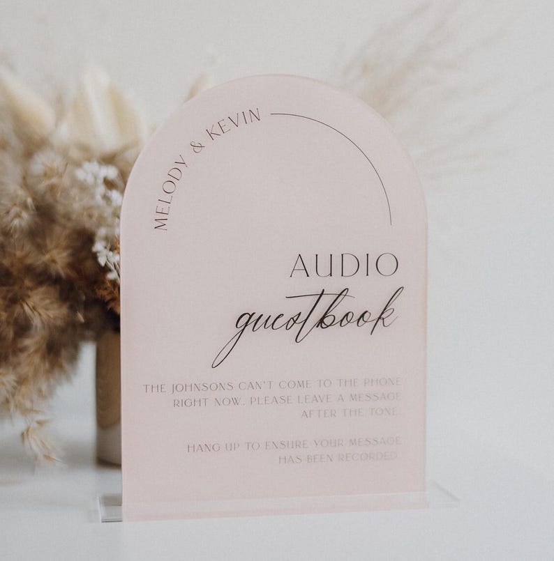 May include: A blush pink acrylic sign with an arched top. The sign reads "MELODY & KEVIN" and "AUDIO guestbook" in elegant script. Below, instructions are printed in a smaller font, guiding guests on leaving a message.