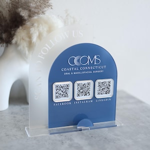 May include: A clear acrylic sign with a blue arched panel featuring the text "COMS Coastal Connecticut Oral & Maxillofacial Surgery." Below are three QR codes with the social media handles Facebook, Instagram, and LinkedIn. The sign reads "SCAN TO FOLLOW US."