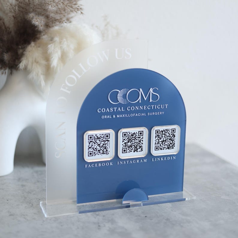 May include: A clear acrylic sign with a blue arched panel featuring the text "COMS Coastal Connecticut Oral & Maxillofacial Surgery." Below are three QR codes with the social media handles Facebook, Instagram, and LinkedIn. The sign reads "SCAN TO FOLLOW US."