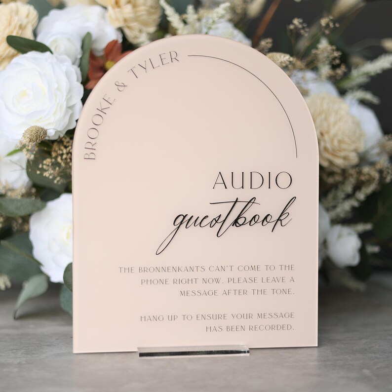 May include: A blush-colored acrylic audio guestbook sign with the names "Brooke & Tyler" at the top. The sign has the words "AUDIO guestbook" in elegant script. Instructions are printed below, with a clear acrylic stand.