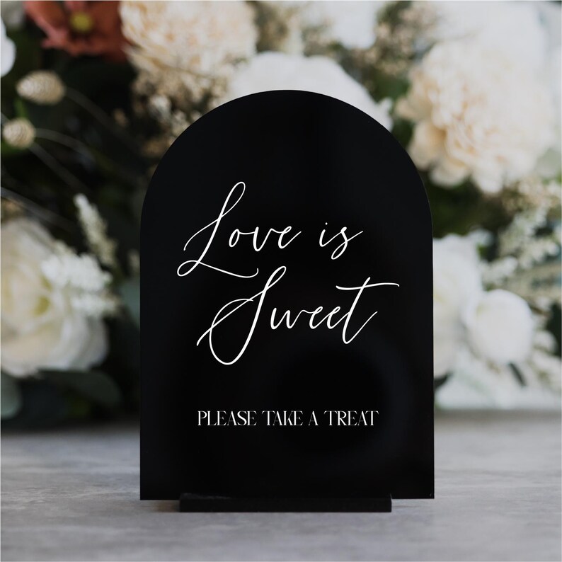 May include: A black, arched sign with the words "Love is Sweet" in elegant white script. Below, the sign reads "PLEASE TAKE A TREAT" in a simple, sans-serif font. The sign is displayed on a gray surface, with a blurred floral background.