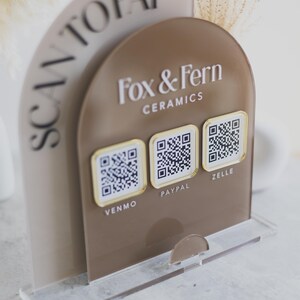 May include: A clear acrylic stand with a brown arched sign that says "Fox & Fern Ceramics." The sign has three QR codes for Venmo, PayPal, and Zelle. The words "SCAN TO PAY" are on a curved panel.