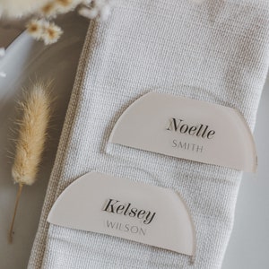 May include: Two white acrylic place cards with rounded edges and a semi-circle shape. The top card reads "Noelle Smith" and the bottom card reads "Kelsey Wilson".