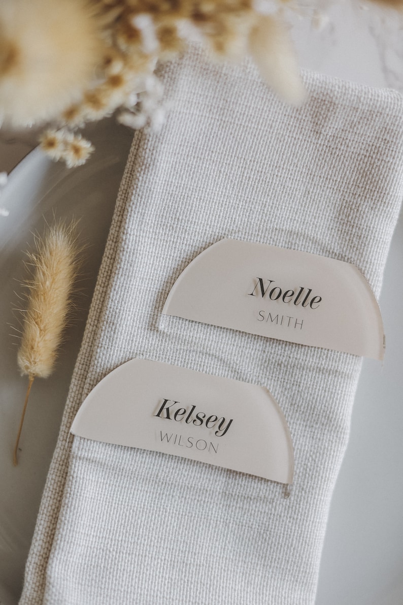 May include: Two white acrylic place cards with rounded edges and a semi-circle shape. The top card reads "Noelle Smith" and the bottom card reads "Kelsey Wilson".