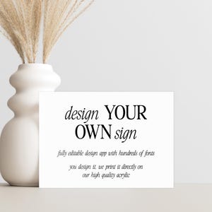 May include: A white rectangular sign with the text "design YOUR OWN sign" in black. The sign also includes text about a design app and printing on acrylic. A white vase with dried plants is to the left.