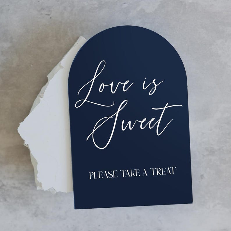 May include: Navy blue arched sign with white script text that reads "Love is Sweet" and "PLEASE TAKE A TREAT". The sign is placed on top of a white paper. Ideal for weddings and events.