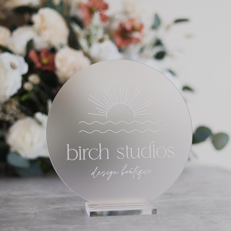 May include: A frosted acrylic sign with a white sun and waves design. The text "birch studios design boutique" is written below the design.