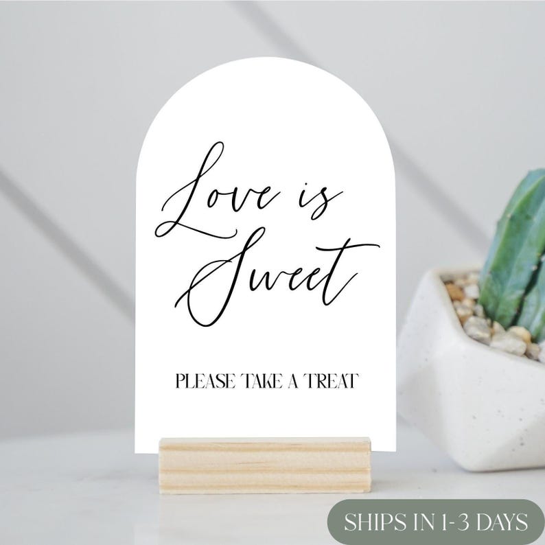 May include: A white sign with an arched top, featuring the text "Love is Sweet" in elegant script, and "PLEASE TAKE A TREAT" in a simple font. The sign is mounted on a small, natural wood base, perfect for weddings or events.