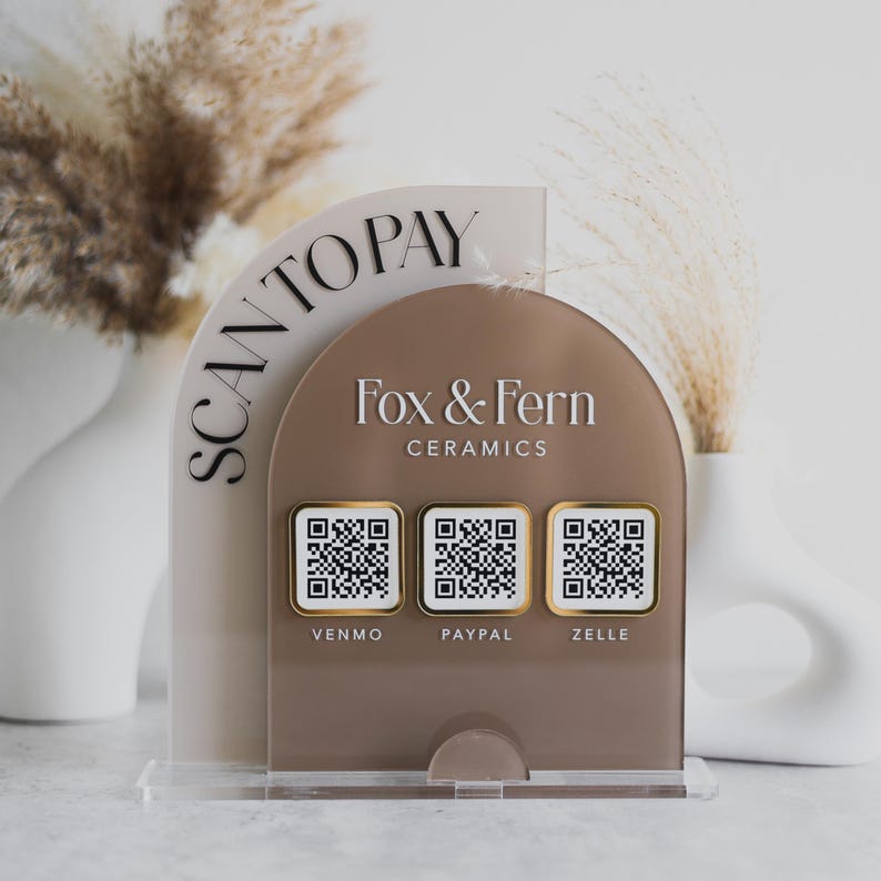 May include: A clear acrylic payment stand with the text "SCAN TO PAY" and the Fox & Fern Ceramics logo. It features three QR codes for Venmo, PayPal, and Zelle. The stand has a neutral colour palette with a clear base.