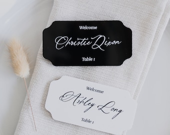May include: Two place cards with a black and white design. The top card is black with white text that reads "Welcome Christie Dixon Table 1". The bottom card is white with black text that reads "Welcome Ashley Long Table 1".