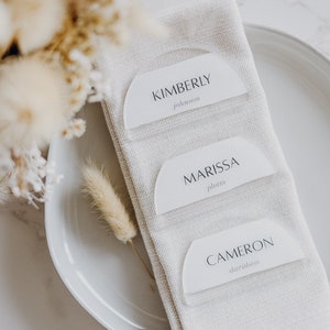 May include: Three white place cards with rounded edges and black text. The cards are arranged vertically on a white linen napkin. The top card reads "KIMBERLY Johnson", the middle card reads "MARISSA Plum", and the bottom card reads "CAMERON Davidson".