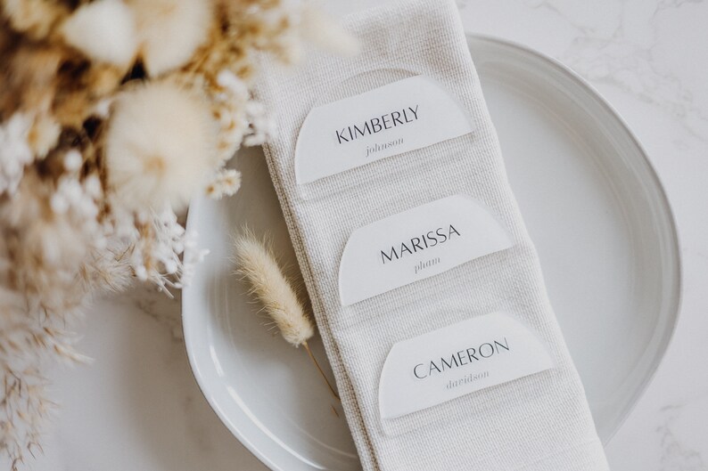 May include: Three white place cards with rounded edges and black text. The cards are arranged vertically on a white linen napkin. The top card reads "KIMBERLY Johnson", the middle card reads "MARISSA Plum", and the bottom card reads "CAMERON Davidson".