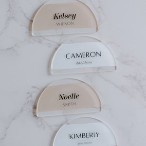 May include: Four acrylic place cards with rounded edges and a half-circle shape. The cards are white with a beige stripe and feature the names Kelsey Wilson, Cameron Davidson, Noelle Smith, and Kimberly Johnson.
