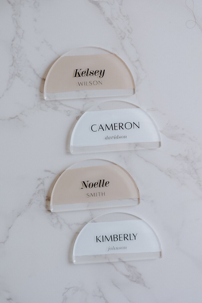 May include: Four acrylic place cards with rounded edges and a half-circle shape. The cards are white with a beige stripe and feature the names Kelsey Wilson, Cameron Davidson, Noelle Smith, and Kimberly Johnson.