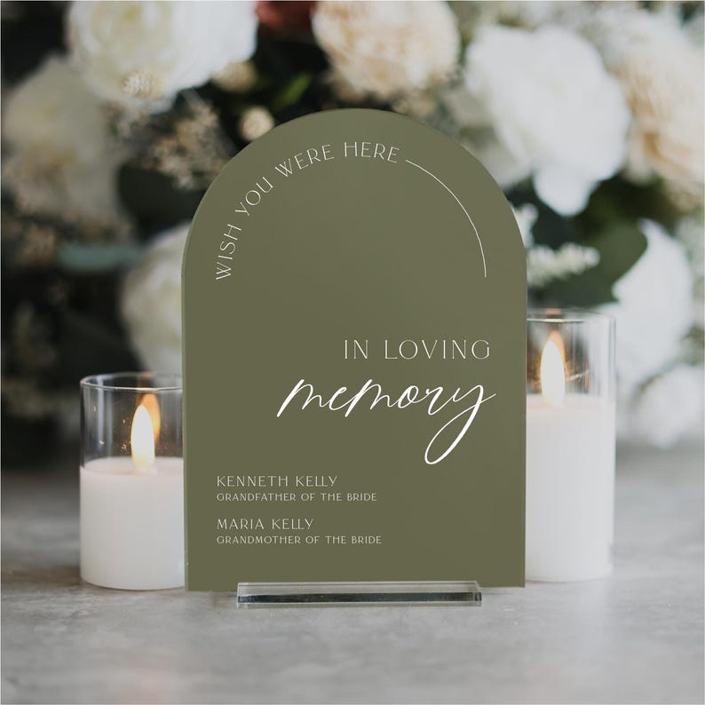 May include: A green acrylic sign with rounded edges and a white inscription that reads "Wish you were here" in a semi-circle at the top. The sign also reads "In loving memory" with the names Kenneth Kelly and Maria Kelly, along with their titles of "Grandfather of the Bride" and "Grandmother of the Bride".