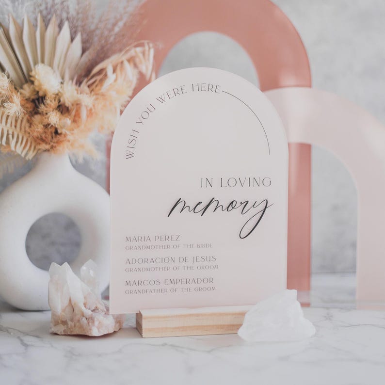 May include: White arch-shaped sign with the text "Wish you were here" in a pink script font. The sign also says "In loving memory" with a list of names and their relation to the bride and groom.