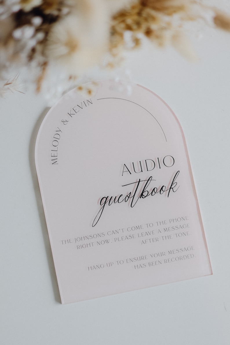 May include: A pink acrylic sign with the words "AUDIO guestbook" in elegant script. The sign includes the names "Melody & Kevin" and instructions for leaving a message. A decorative wedding or event sign.