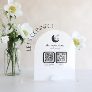 May include: A white acrylic sign featuring the text "LET'S CONNECT" and the logo for "the moonstone candle company." Two QR codes are below the logo, labelled "INSTAGRAM" and "SHOP." A vase of white flowers is to the left.