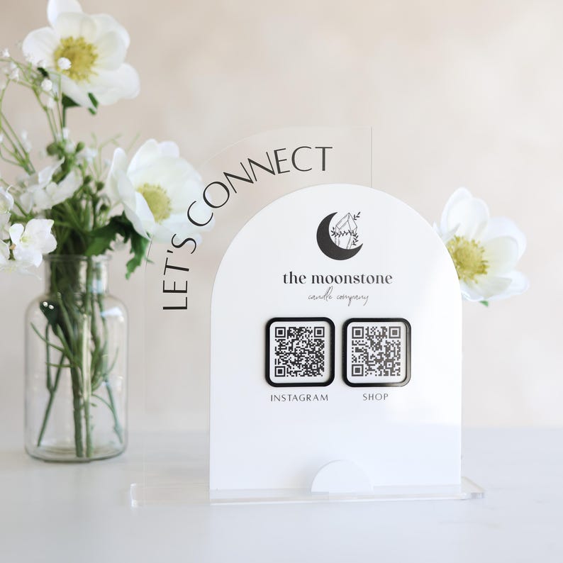 May include: A white acrylic sign featuring the text "LET'S CONNECT" and the logo for "the moonstone candle company." Two QR codes are below the logo, labelled "INSTAGRAM" and "SHOP." A vase of white flowers is to the left.