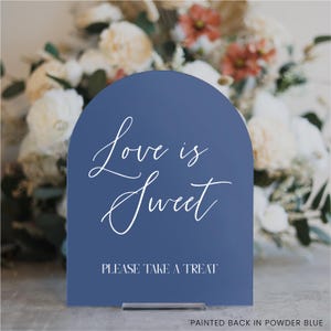 May include: A navy blue arched sign with the words "Love is Sweet" in elegant white script. Below, it reads "PLEASE TAKE A TREAT". The sign is displayed on a clear stand, with a blurred floral arrangement in the background.