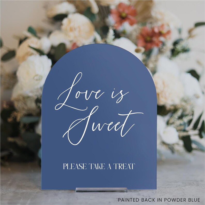 May include: A navy blue arched sign with the words "Love is Sweet" in elegant white script. Below, it reads "PLEASE TAKE A TREAT". The sign is displayed on a clear stand, with a blurred floral arrangement in the background.