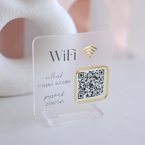 May include: Clear acrylic WiFi sign with gold accents. The sign displays the word "WiFi" and a gold wireless symbol. It includes a QR code and the network name "Coppice & Crafts" with the password "23567345".