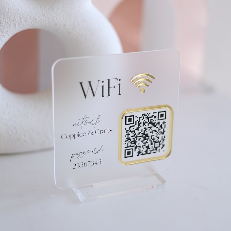 May include: Clear acrylic WiFi sign with gold accents. The sign displays the word "WiFi" and a gold wireless symbol. It includes a QR code and the network name "Coppice & Crafts" with the password "23567345".