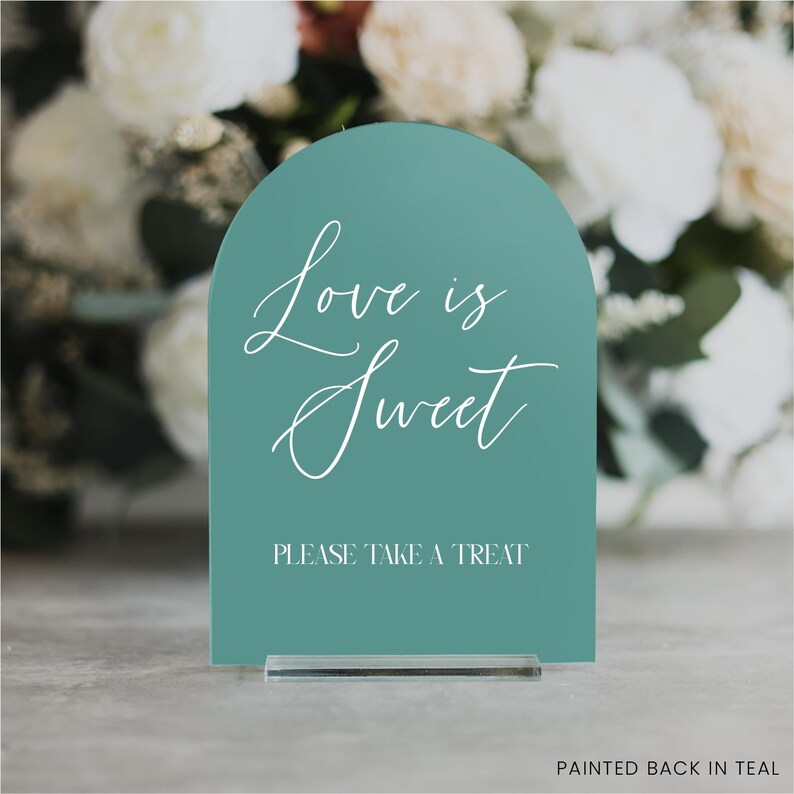 May include: Teal-colored sign with an arched top, featuring the text "Love is Sweet" in elegant script and "PLEASE TAKE A TREAT" in a simple font. The sign is displayed on a clear acrylic base.