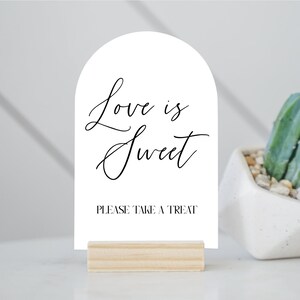May include: A white sign with an arched top, featuring the text "Love is Sweet" in elegant script, and "PLEASE TAKE A TREAT" in a simple font. The sign is held by a small, natural wood base.
