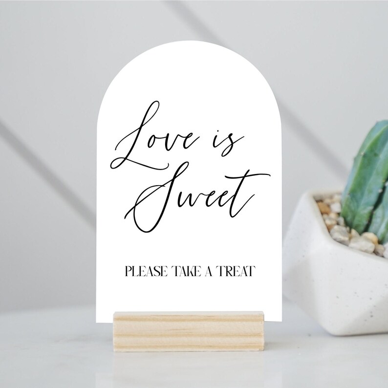 May include: A white sign with an arched top, featuring the text "Love is Sweet" in elegant script, and "PLEASE TAKE A TREAT" in a simple font. The sign is held by a small, natural wood base.