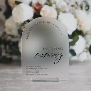 May include: A frosted acrylic arch sign with the text "Wish you were here" and "In loving memory". The sign also includes the names Kenneth Kelly, Grandfather of the Bride, and Maria Kelly, Grandmother of the Bride.