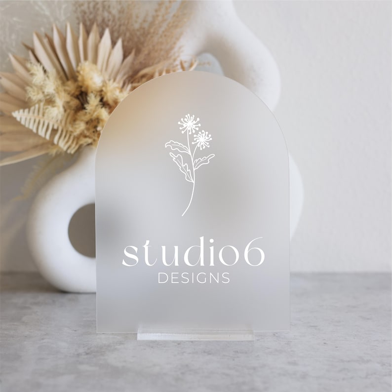 Może przedstawiać: Biały, matowy znak akrylowy z zaokrąglonym g&oacute;rnym brzegiem. Znak zawiera prosty rysunek kwiatu i tekst "studio6 DESIGNS".