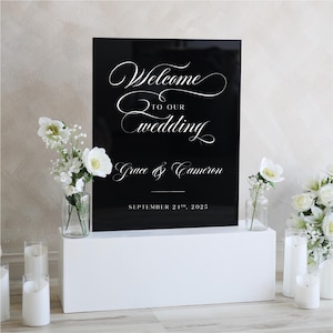 May include: A black wedding sign with white calligraphy that reads "Welcome to our Wedding" followed by the names "Grace & Cameron" and the date "September 24th, 2025". The sign is on a white pedestal with floral arrangements.