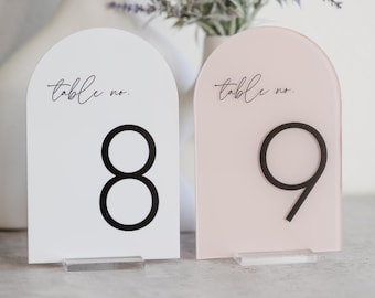 May include: Two acrylic table number signs with rounded tops. The sign on the left is white with black text and the number 8. The sign on the right is pink with black text and the number 9. Both signs have the text "table no."