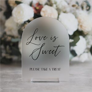 May include: Frosted acrylic sign with an arched top. The sign reads "Love is Sweet" in elegant script, with "PLEASE TAKE A TREAT" below. The sign is displayed on a clear acrylic base, perfect for weddings or events.