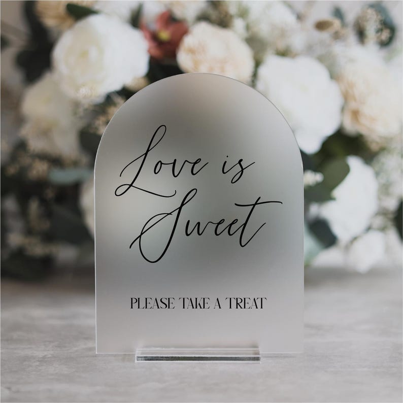 May include: Frosted acrylic sign with an arched top. The sign reads "Love is Sweet" in elegant script, with "PLEASE TAKE A TREAT" below. The sign is displayed on a clear acrylic base, perfect for weddings or events.