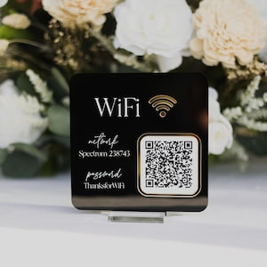 May include: A black square WiFi sign with gold text and a QR code. The sign displays the word "WiFi", network name, and password. The sign is displayed on a clear acrylic stand, with a floral arrangement in the background.