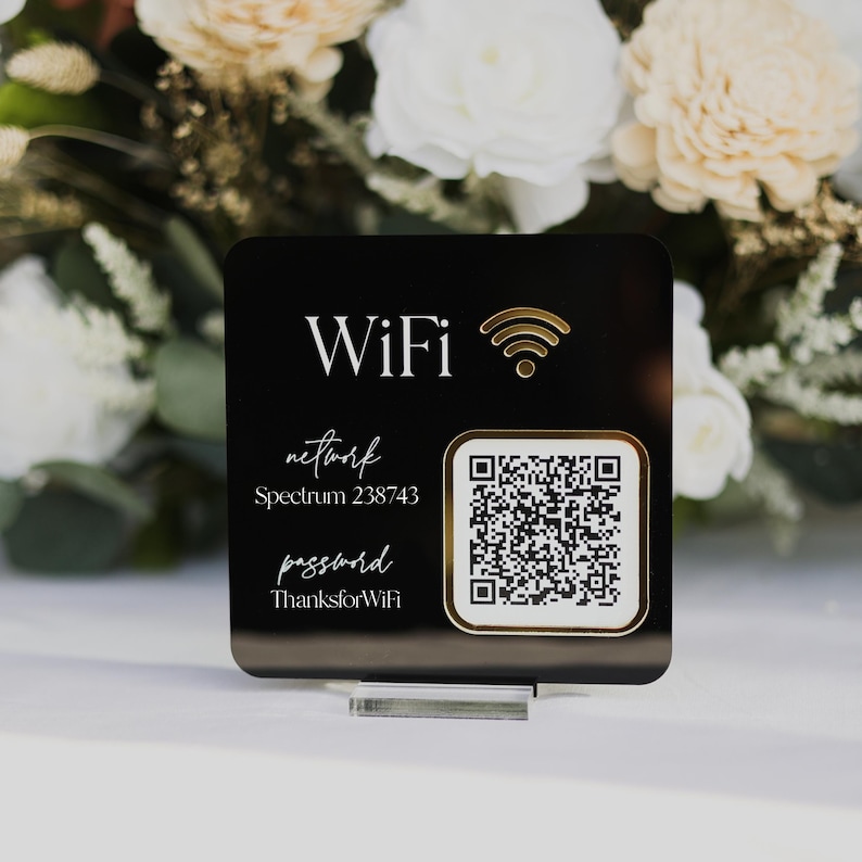May include: A black square WiFi sign with gold text and a QR code. The sign displays the word "WiFi", network name, and password. The sign is displayed on a clear acrylic stand, with a floral arrangement in the background.