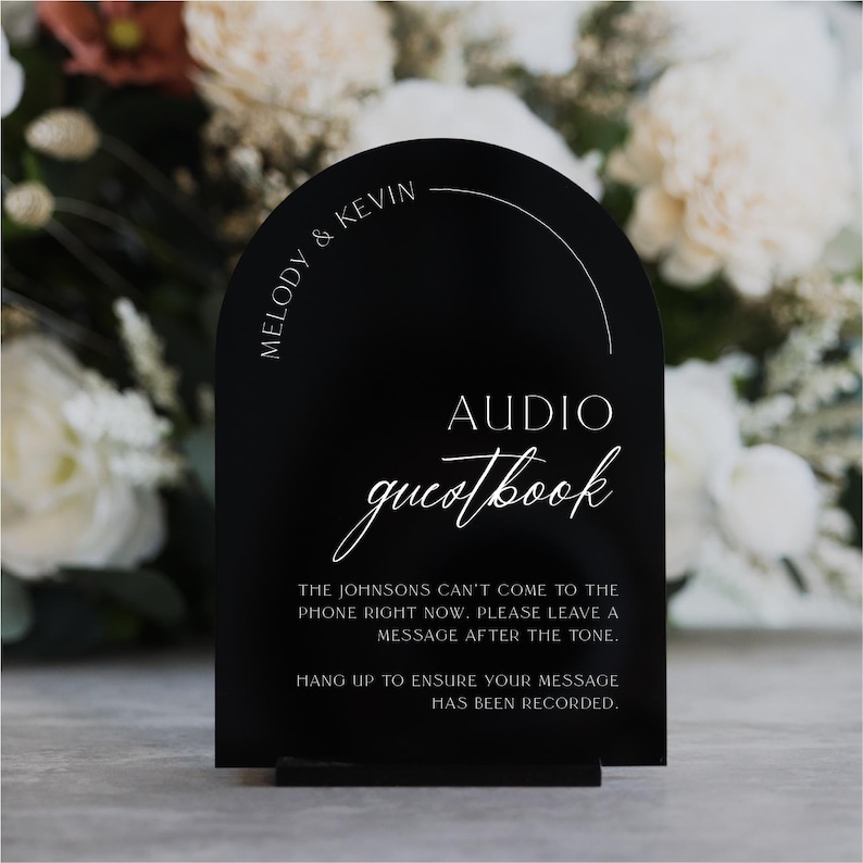 May include: A black acrylic wedding sign with an arched top. The sign reads "AUDIO guestbook" in elegant script. The names "Melody & Kevin" are printed at the top. Instructions for leaving a message are printed below.