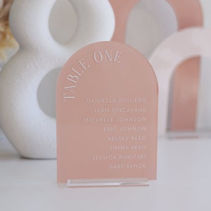 May include: A blush pink acrylic table number sign with a rounded arch top. The sign says "TABLE ONE" in white lettering and lists the names of guests: Daniella Diguero, Ivan Sincavage, Michelle Johnson, Eric Johnon, Kelsey Reed, Timmy Reed, Jessica Rokitski, and Gabe Benza.