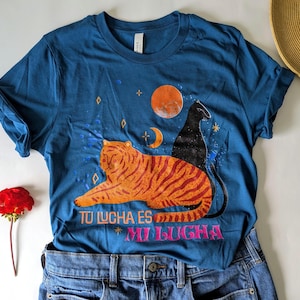 May include: Teal t-shirt with a graphic of an orange tiger, a black cat, a crescent moon, and a red sun. The text "TU LUCHA ES MI LUCHA" is printed in pink. The shirt is folded on top of blue jeans.