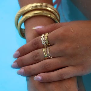 May include: Gold-toned jewelry set against a blue background. The image features a thick, textured bracelet and multiple rings. One ring has a floral design, and another has a unique, curved shape. The jewelry is displayed on a hand.