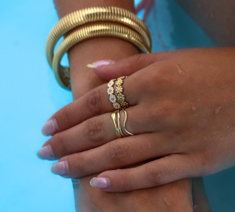 May include: Gold-toned jewelry set against a blue background. The image features a thick, textured bracelet and multiple rings. One ring has a floral design, and another has a unique, curved shape. The jewelry is displayed on a hand.