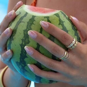 May include: A halved watermelon with green and dark green striped rind and red flesh is held by hands with long, manicured nails. The hands wear gold rings, one with a floral design. A gold bracelet is visible.